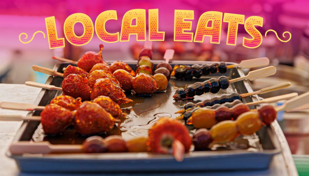 Colorful skewers of candied fruit are displayed on a tray under a bright "Local Eats" sign.