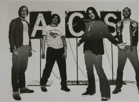 Four men stand in front of large letters spelling "ACS"; the image is in black and white, and each person faces the camera with casual or open body language.