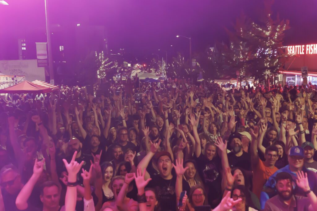 Large crowd at an outdoor event at night, many people raising their hands, with a building reading “Seattle Fish” illuminated in the background.