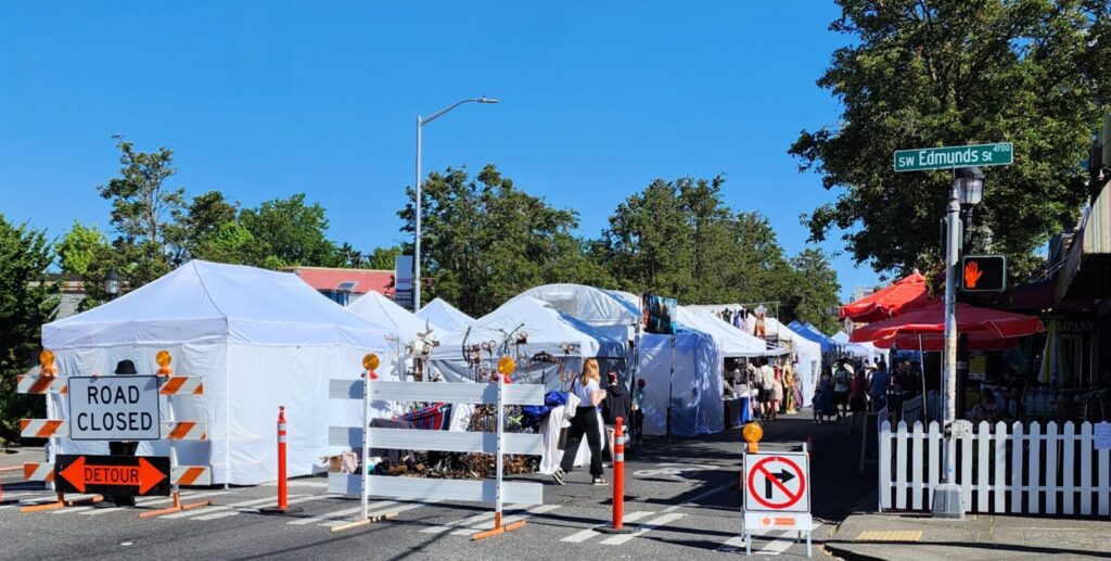 Outdoor street market with white tents, vendor stalls, and people walking. "Road Closed" and detour signs block vehicle access. Visible street sign reads "SW Edmunds St.