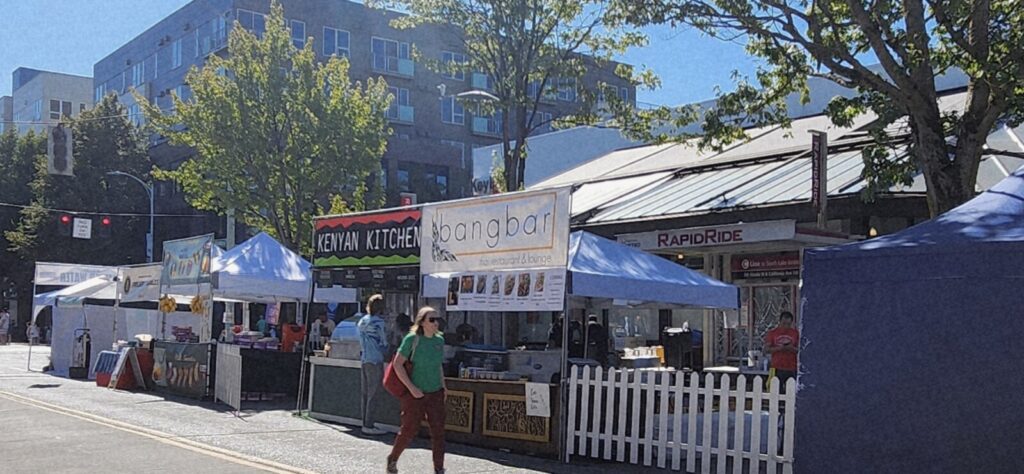 Outdoor street market with food vendor tents, including signs for Keyaki Kitchen and Bangbar, set up along a city sidewalk on a sunny day. Several people are walking by.