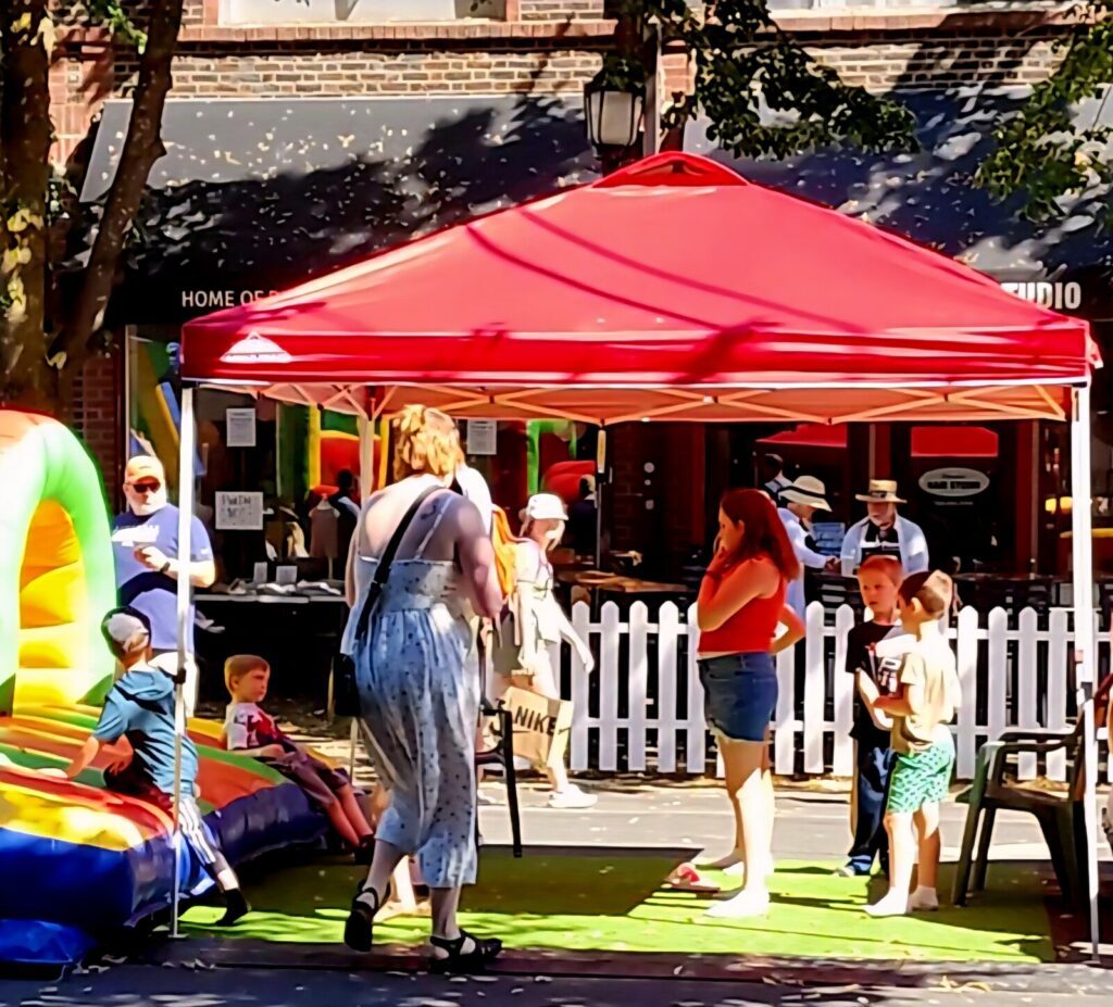Children and adults gather under a red canopy next to an inflatable play structure at an outdoor event on a sunny day.