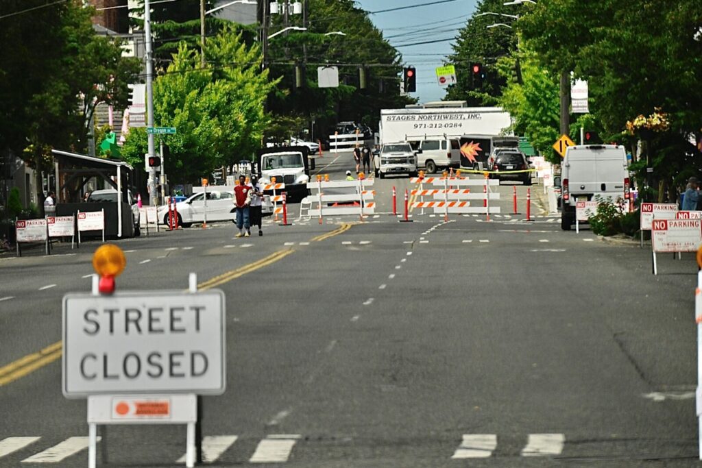 A city street is closed with barricades and signs, blocking vehicle access. People and vehicles are seen in the background near the closure.