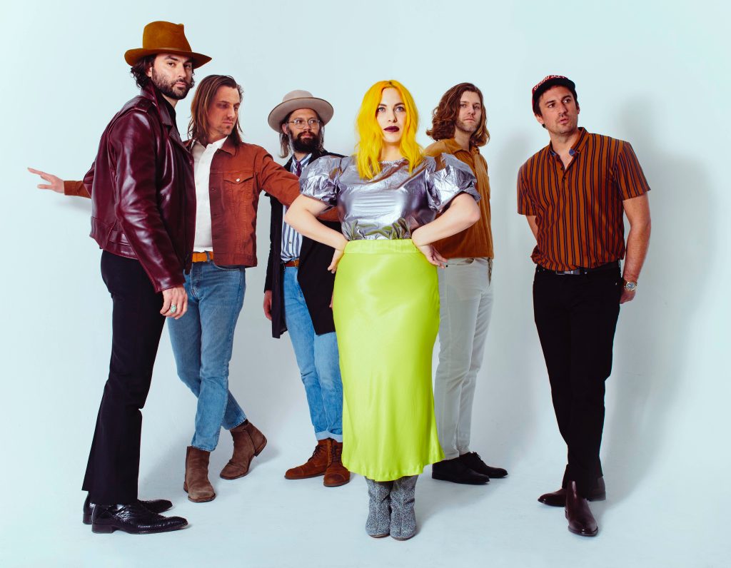 Six people stand posing against a white background; five men in casual outfits and a woman in a metallic top and bright yellow skirt stands at the center.
