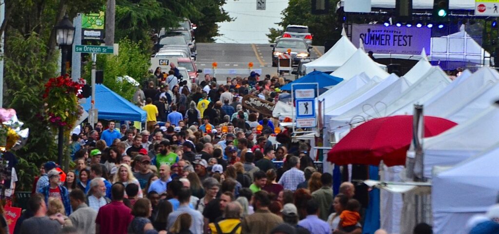 A crowd walks through a street lined with vendor tents at an outdoor festival. Some people wear bright shirts, and there are trees and street signs visible in the background.