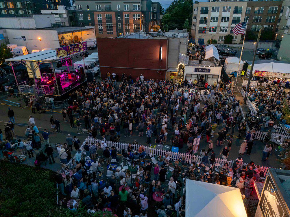 A large crowd gathers at an outdoor festival with a stage, food booths, and American flags, surrounded by multi-story buildings in the evening.