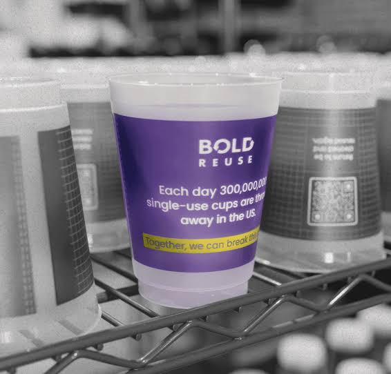 Plastic cup on a shelf with a label stating that 300 million single-use cups are thrown away daily in the US; features the text "BOLD REUSE" and a QR code.