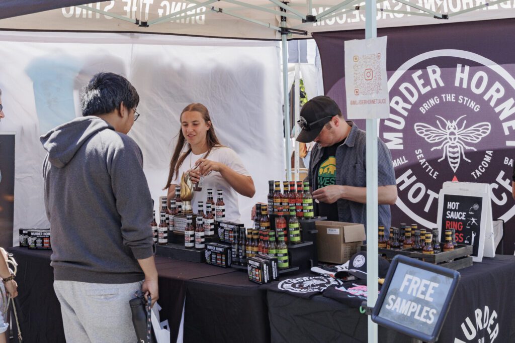 Two vendors stand behind a table displaying jars of hot sauce at an outdoor market booth. A person stands in front, and a "Free Samples" sign is visible on the table.