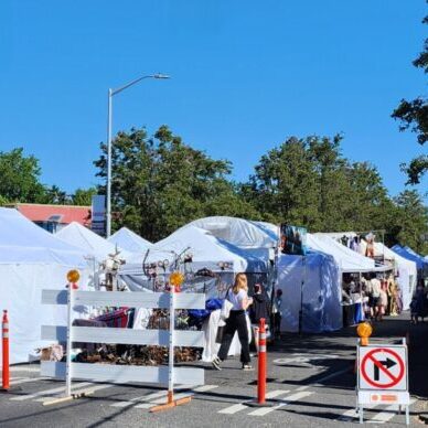 Outdoor street market with white tents, vendor stalls, and people walking. "Road Closed" and detour signs block vehicle access. Visible street sign reads "SW Edmunds St.