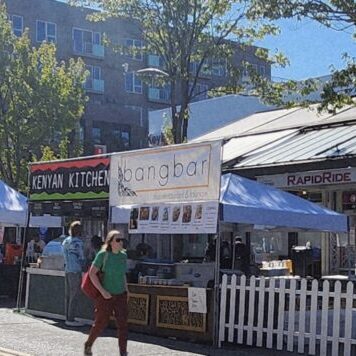 Outdoor street market with food vendor tents, including signs for Keyaki Kitchen and Bangbar, set up along a city sidewalk on a sunny day. Several people are walking by.