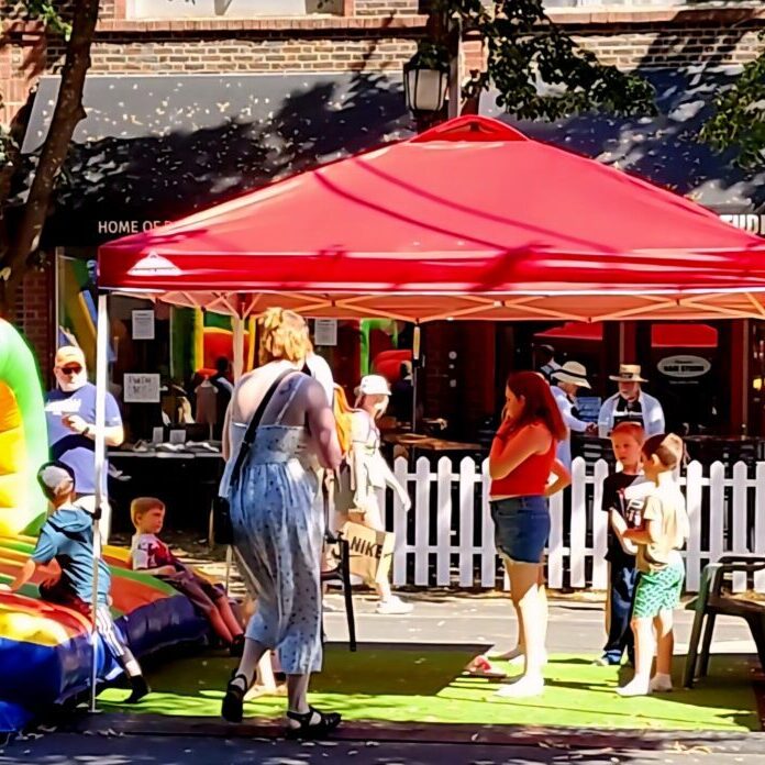 Children and adults gather under a red canopy next to an inflatable play structure at an outdoor event on a sunny day.