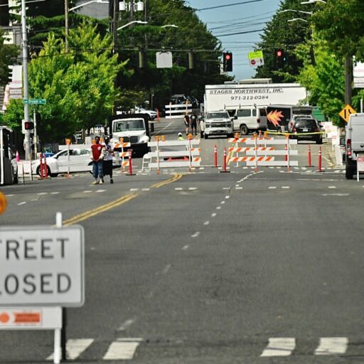 A city street is closed with barricades and signs, blocking vehicle access. People and vehicles are seen in the background near the closure.