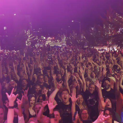 Large crowd at an outdoor event at night, many people raising their hands, with a building reading “Seattle Fish” illuminated in the background.