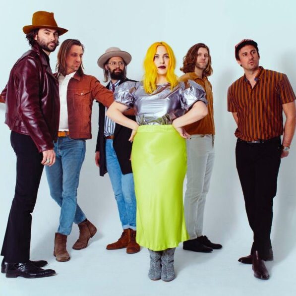Six people stand posing against a white background; five men in casual outfits and a woman in a metallic top and bright yellow skirt stands at the center.