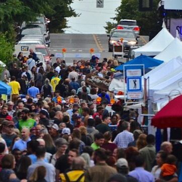 A crowd walks through a street lined with vendor tents at an outdoor festival. Some people wear bright shirts, and there are trees and street signs visible in the background.