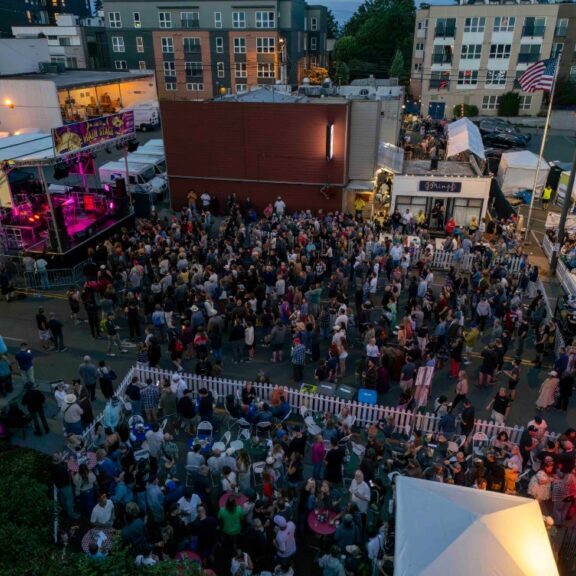 A large crowd gathers at an outdoor festival with a stage, food booths, and American flags, surrounded by multi-story buildings in the evening.