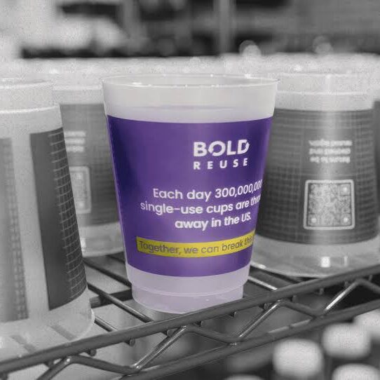 Plastic cup on a shelf with a label stating that 300 million single-use cups are thrown away daily in the US; features the text "BOLD REUSE" and a QR code.